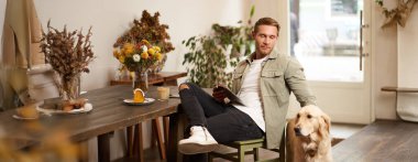 Yakışıklı genç adamın portresi köpeğiyle kafede oturuyor Golden Retriever 'ı okşuyor, tabletten haberler okuyor, bir fincan kahveyle rahatlıyor..