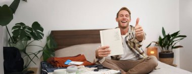 Neşeli genç bir adam, kontrol listesi olan bir turist, notlarını defterde gösteriyor, baş parmaklarını kaldırıyor, bavullarını topluyor, tatile çıkıyor, yurt dışına çıkıyor ve valizlerini hazırlıyor. Boşluğu kopyala