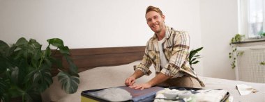 Genç yakışıklı sakallı adamın portresi, tatile çıkan bir turist, giysilerini bavula bağlıyor, tatile çıkıyor, iş gezisine çıkıyor, yatak odasında oturuyor..