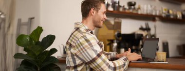 Yakışıklı gülümseyen bir adamın portresi, bir kafede dizüstü bilgisayarla oturmak, birlikte çalışma alanından uzaktan çalışmak, kahve içmek, kamu alanından serbest çalışmak..