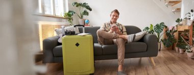 Yakışıklı genç adam kanepede bavuluyla oturuyor, elinde akıllı telefon, kitap pansiyonu ya da tatil tatili var, cep telefonuyla sohbet ediyor. Turizm ve seyahat kavramı