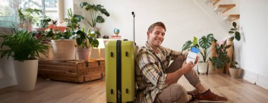 Erkek modelin bavullu portresi, yerde oturuyor, uçak biletleri ve pasaport tutuyor, yurtdışına gidiyor, havaalanına gitmek için taksi bekliyor..