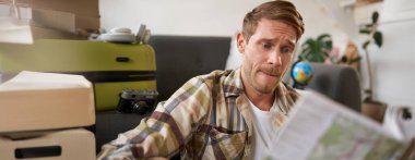 Seyahat haritasına kafası karışmış bir adamın portresi, rotayı seçmek, tatile gitmek, tatile hazırlanmak, oturma odasında bavulla oturmak..