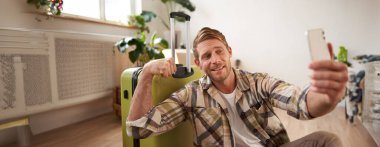 Yakışıklı, gülümseyen genç bir adamın tatile çıkması, tatille ilgili internette paylaşım yapması, akıllı telefondan bavulla selfie çekmesi, kaslarını gösteriyor..