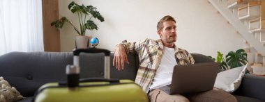 Mutlu genç adamın portresi, turist seyahat acentesine rezervasyon yaptırıyor, dizüstü bilgisayara bakıyor, tatile gitmek için bavulunu hazırlıyor..