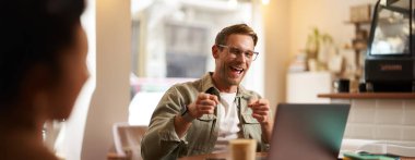 Gözlüklü mutlu genç adam, kafede dizüstü bilgisayarla oturuyor, kahve içiyor, kamerayı işaret ediyor ve neşeyle gülümsüyor..