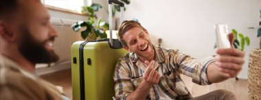 Elinde bavuluyla gülümseyen genç adam, akıllı telefondan selfie çekiyor, sosyal medyada tatil turu hakkında blog yazıları yazıyor, cep telefonu kullanıyor. Turizm ve gezgin insanlar konsepti