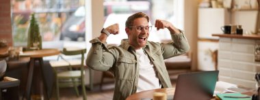 Gözlüklü mutlu bir gencin portresi neşe içinde kafede oturuyor, laptopta spor maçı izliyor, kutlama yapıyor, kaslarını gösteriyor ve gülüyor..