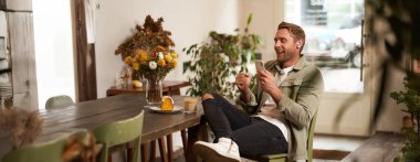 Kafede cep telefonuyla oturan, kablosuz kulaklık takan kahve dükkanında dinlenirken müzik ya da podcast dinleyen mutlu genç adamın portresi..