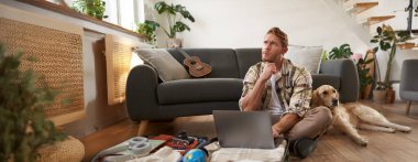 Genç bir adamın portresi, gezgin bir köpekle tatile çıkıyor, bavulunu topluyor, laptopuyla oturuyor, düşünüyor, online rezervasyon yapıyor..