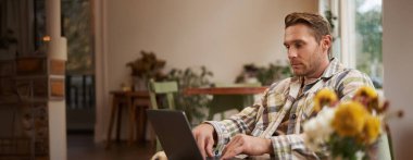 Yaşam tarzı ve iş yeri konsepti. Günlük giysiler içinde genç yakışıklı bir adam, kafeteryada dizüstü bilgisayarla oturuyor, internetten çalışıyor, kahve dükkanında uzaktan çalışıyor..