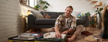 Genç bir adamın görüntüsü, gezgin, açık bavulla yerde oturuyor, seyahat için giysilerini topluyor, tatile çıkıyor. Turizm konsepti