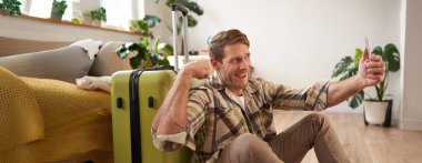 Komik yakışıklı adam, turist kasları kasıyor, tatile çıkıyor, bavulla oturuyor, akıllı telefondan selfie çekiyor. Turizm ve seyahat kavramı