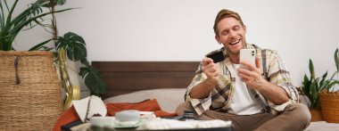 Neşeli turist, kredi kartı ve cep telefonu olan adam, tatil biletlerini ödeyen, bavulla yatakta oturan, toplanan ve tatile çıkan adam..