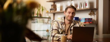 Yakışıklı genç dijital göçebe portresi, kafede dizüstü bilgisayarla çalışan, online projesinden memnun ve mutlu görünen, birlikte çalıştığı kahve dükkanında oturan bir adam..