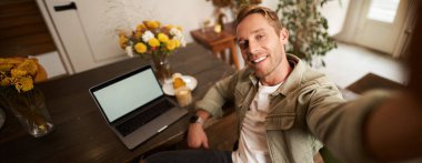 Kafede dizüstü bilgisayarla fotoğraf çeken ve kameraya gülümseyen yakışıklı bir gencin selfie portresi..