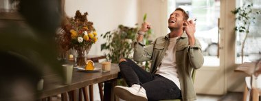 Rahatlamış, mutlu genç bir adamın portresi, mutlu haberler dinliyor ya da kulaklıkla podcast yapıyor, kahve dükkanında oturuyor, akıllı telefonu tutuyor, elini kaldırıyor ve heyecandan gülümsüyor, kafede vakit geçiriyor..