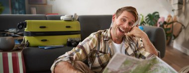 Yakışıklı, gülümseyen genç bir adamın portresi, turist gösterileri bana jest, telefon eli, seyahat haritası, tatile gitmek için davetiye, valiz.