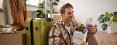 Elinde bavuluyla gülümseyen yakışıklı bir adamın portresi, akıllı telefona bakıyor, seyahat bileti ve pasaport tutuyor. Turizm ve seyahat kavramı