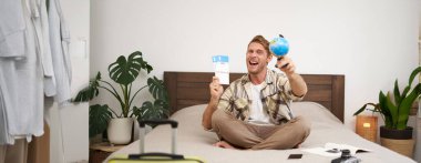 Heyecanlı, yakışıklı genç adam yatağa oturur, dünyayı ve biletleri gösterir, bavulunu hazırlar, tatile çıkar, mutlu bir şekilde tatile çıkar. Turizm konsepti