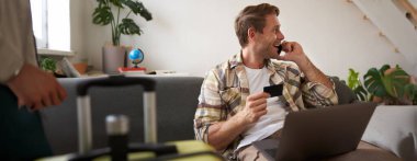 Kredi kartı tutan ve dizüstü bilgisayar kullanan bir adamın resmi. Tatil için bavul ve kamera hazırlamış. Turizm konsepti