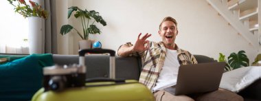 Mutlu adam internetten tatil rezervasyonu yapıyor, seyahat acentesinin internet sitesinden tatil parası ödüyor, internetten bilet alıyor, oturma odasında bavuluyla oturuyor..