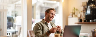 Yakışıklı, gülümseyen genç adamın portresi, çevrimiçi öğretmen, kafede uzaktan çalışan iş adamı, dizüstü bilgisayara el sallayan, görüntülü sohbete ya da buluşmaya bağlanan.