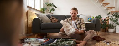 Seyahat ve turizm konsepti. Mutlu genç adam, bagajı açık bir şekilde yerde oturuyor, tatil için bavulunu hazırlıyor, tatile çıkıyor, kıyafetlerini bir çantaya koyuyor..