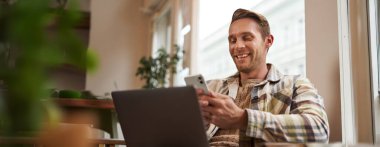 Bilgisayarlı, yakışıklı, gülümseyen bir adamın portresi. Kafe ziyaretçisi sandalyede oturuyor, akıllı telefon kullanıyor, mutlu görünüyor, arkadaşlarıyla mesajlaşıyor ve kahve içiyor..