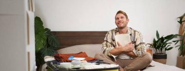 Düşünceli genç bir adamın portresi, tatilini planlayan bir turist, açık bavulla yatakta oturuyor, notlar alıyor, not defterine bir şeyler yazıyor..