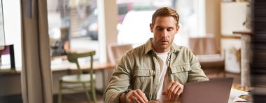 Kafede dizüstü bilgisayarla oturup gözlük takan işine odaklanmış genç bir adamın görüntüsü..