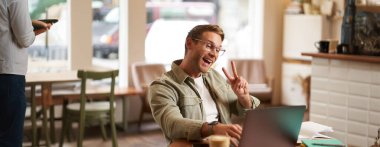 Gözlüklü mutlu adam, kafede oturuyor, diz üstü bilgisayarda barış işareti gösteriyor, görüntülü sohbetler, çevrimiçi toplantılara bağlanıyor..