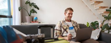 Yakışıklı bir gencin portresi, iki bileti ve pasaportu var, dizüstü bilgisayara bağlanıyor, otel rezervasyonu yapıyor ve tatilini planlıyor, oturma odasında oturuyor..