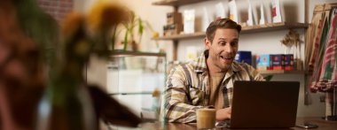 Yakışıklı genç dijital göçebe portresi, kafede dizüstü bilgisayarla çalışan, online projesinden memnun ve mutlu görünen, birlikte çalıştığı kahve dükkanında oturan bir adam..