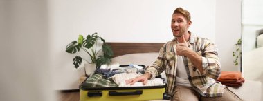 Çekici genç adam gülümsüyor, başparmağını kaldırıyor, valizin içindeki kıyafetlerini gösteriyor, tatile hazırlanıyor, seyahat acentesiyle seyahat ediyor. Yaşam tarzı ve turizm konsepti