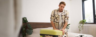 Yatakta oturan bir adamın portresi, valizi kapatmaya çabalayan, tatil için hazırlanan, tatile çıkan. Turizm ve seyahat kavramı