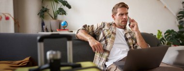 Kredi kartlı genç bir adamın portresi, oturuyor ve dizüstü bilgisayar kullanıyor, bir telefon görüşmesi yapıyor, otel ya da bilet ayırtıyor, telefonla satın alımını onaylıyor, yanında bavulu var..