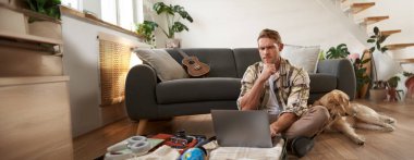 Genç bir adamın resmi, tatile çıkan bir turist, oturma odasında bir köpekle oturuyor, dizüstü bilgisayara bakıyor, bilgisayardaki bir uçağa giriş yapıyor. Turizm ve seyahat kavramı
