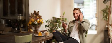 Kafedeki yakışıklı adam, kablosuz kulaklıkla müzik dinliyor, akıllı telefon çalma listesindeki favori şarkının tadını çıkarmak için kafeye bağlanıyor, oturup cappuccino içiyor..