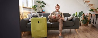 Adamın portresi oturma odasında cep telefonuyla oturuyor, bavulunu hazırlıyor, akıllı telefon uygulamasında sohbet ediyor, tatil yerlerine bakıyor, tatile çıkıyor..