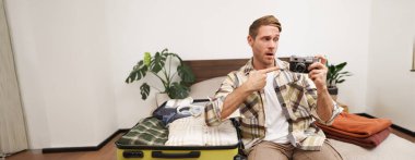 Yakışıklı mutlu adamın portresi, analog kamerasını tutuyor, açık bavulun yanında oturuyor, tatil için eşyalarını topluyor, yurt dışına seyahat ediyor, tatile hazırlanıyor..