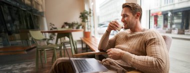 Yakışıklı modern adamın portresi, kafeteryada dizüstü bilgisayarla oturuyor, mutlu yüzle ekrana bakıyor, cappuccino içiyor, kafede dinleniyor ya da çalışıyor..