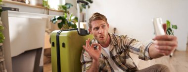 Heyecanlı genç adam bavulunu gösteriyor, cep telefonuyla selfie çekiyor, bavulla poz veriyor, tatile çıkıyor. Boşluğu kopyala