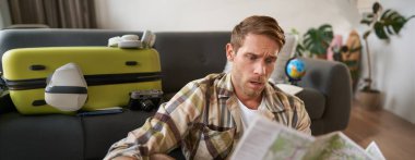 Endişeli, kafası karışmış, haritaya bakan, rotayı incelerken kaşlarını çatan, bavulla oturan, tatile çıkan bir adamın portresi..