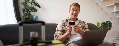 Neşeli bir adamın portresi, tatile çıkıyor, yurtdışına seyahat planları yapıyor, dizüstü bilgisayarı açık, oturma odasında bavuluyla oturuyor, bir otel rezervasyonu yaptırıyor..