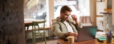 Kafede çalışan bir adamın portresi, kameraya şaşırmış bir şekilde bakmak, dizüstü bilgisayar ekranına bakmak için gözlüklerini çıkarmak, bilgisayarında bir sorusu var. Uzak görevler konsepti.