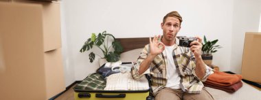 Genç yakışıklı bir adamın portresi, seyahat fotoğrafçısı, tamam işareti, yatakta kamerayla oturuyor, bavulları bavula dolduruyor, tatile çıkıyor ya da yurtdışına tatile çıkıyor..