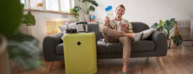 Genç turistin portresi, koltukta bavullu bir adam, tatile hazırlanıyor, akıllı telefonla oturuyor, cep telefonu uygulamasından otel ve uçak biletleri alıyor. Turizm ve seyahat kavramı