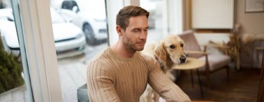 Yakışıklı genç bir adam, dijital göçebe köpeğiyle birlikte evcil hayvan dostu bir kafede oturuyor, bir Golden Retriever onu izlerken dizüstü bilgisayarda çalışıyor..