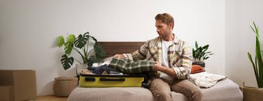 Yolculuk için bavulunu toplayan genç adamın portresi, tatile gitmek, yatağa oturmak ve giysilerini bavula koymak, uçuşu için hazırlanmak..